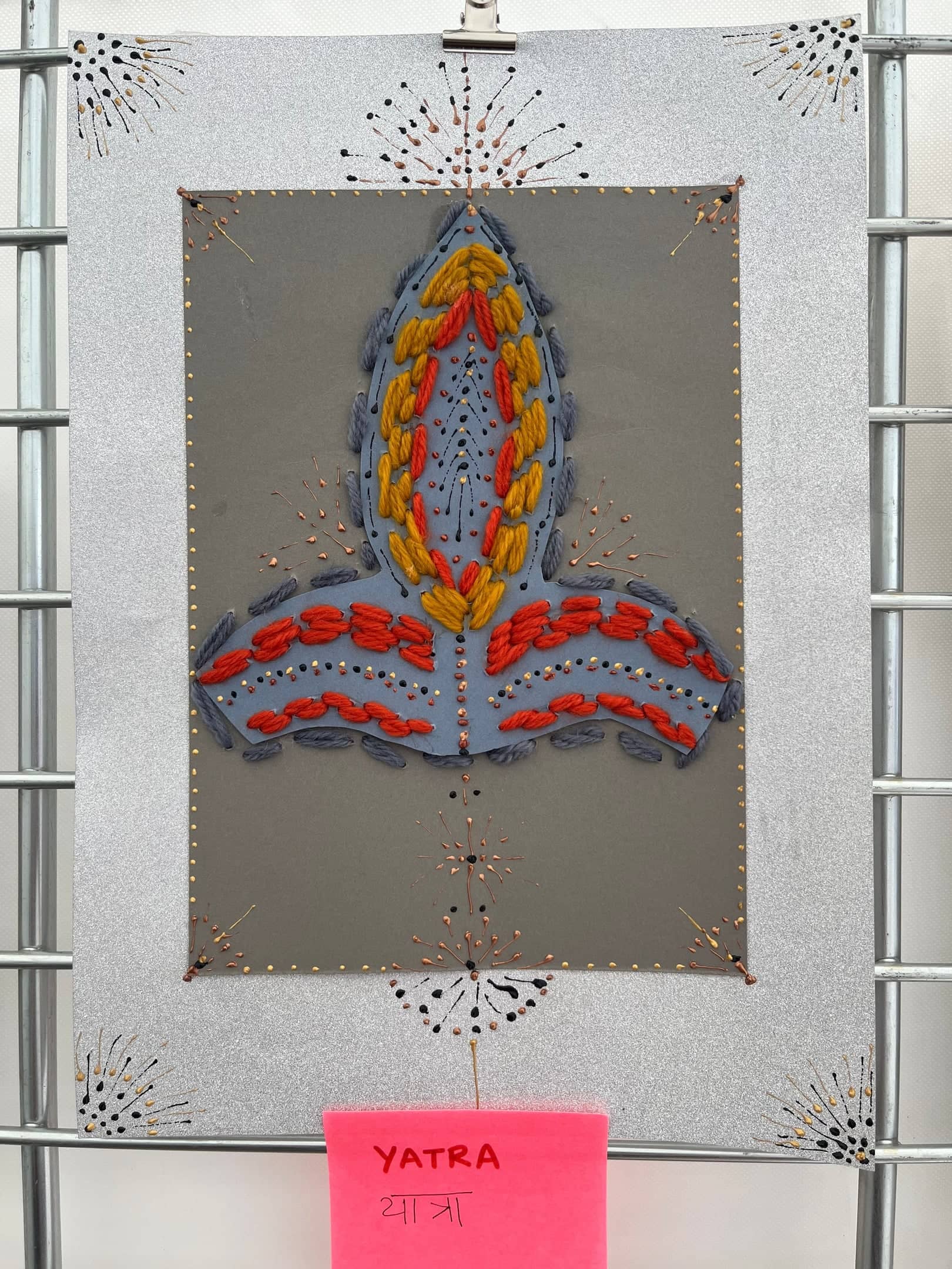 Yatra, av Anujalika. En flamliknande form broderad i orange, gult, rött och grått ullgarn på grå kartong, omgiven av linjer av små mörka stygn som strålar utåt; en rosa etikett lyder Yatra.