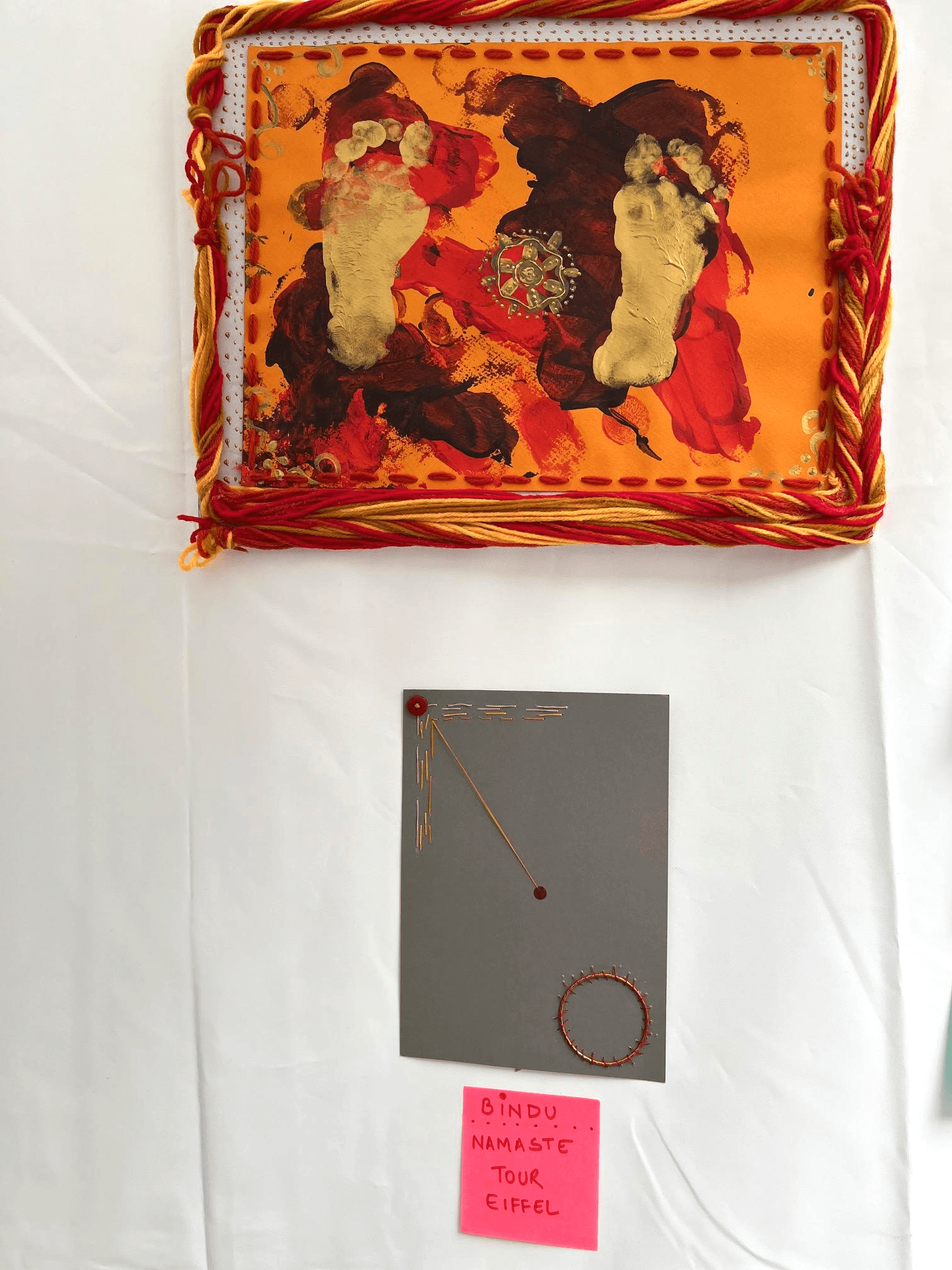 Bindu, av Anujalika. En orange textil med flätad röd och gul bård hänger ovanför en liten grå kartong märkt med en enda röd punkt och en röd cirkel.