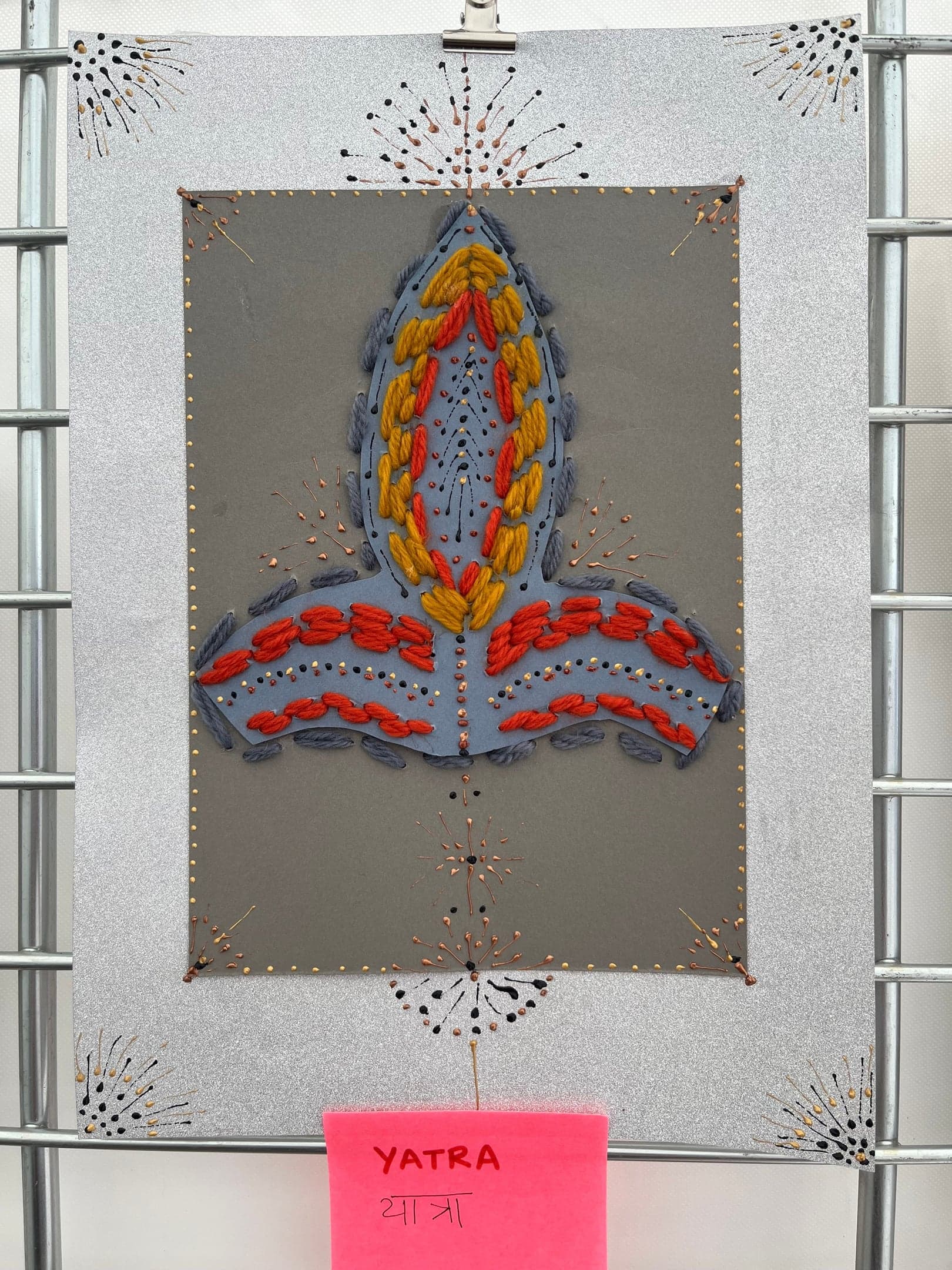 Yatra, by Anujalika. A flame-like form stitched in orange, yellow, red and grey wool on grey card, with lines of small dark stitches radiating outward; a pink label reads Yatra.
