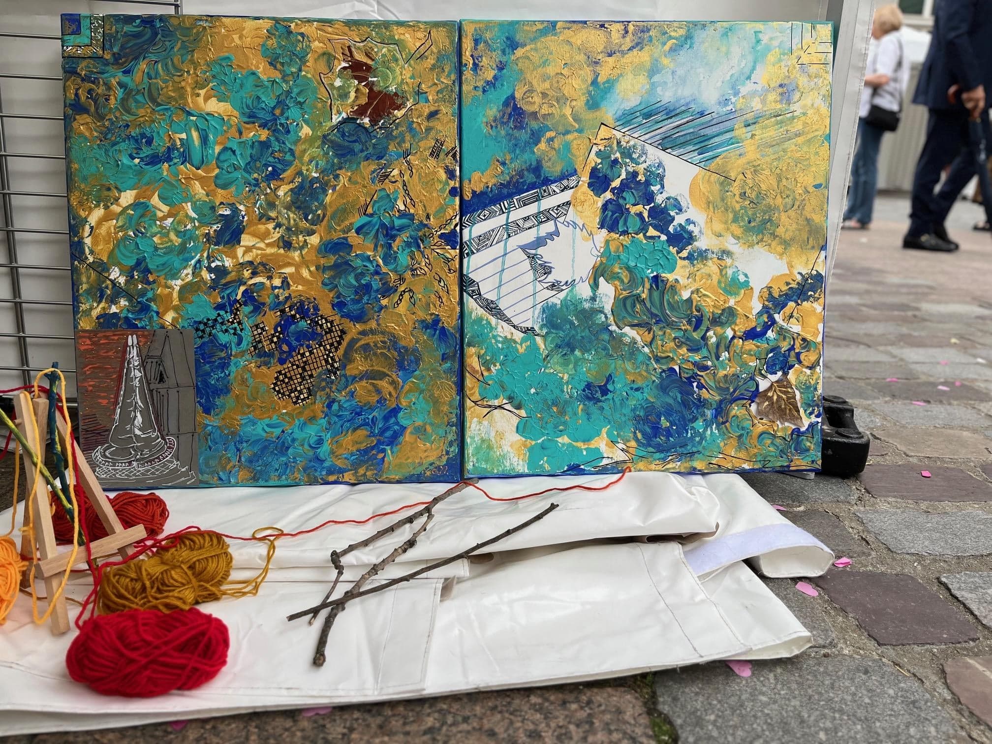 Namaste Tour Eiffel, Mixed media, 2021, by Anujalika. Two canvases side by side, poured in blue, turquoise and gold, with a black line drawing of the Eiffel Tower crossing the seam; skeins of red, orange and mustard wool lie in front.